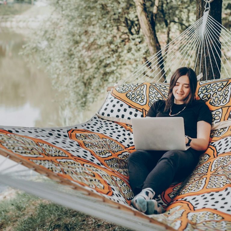 Persona sonriente sentada en una hamaca, usando una laptop junto a un río.